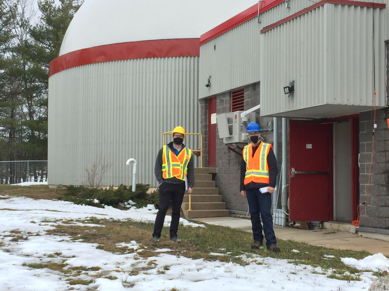 Two people wearing hard hats and safety vests outside a facility with a large domed tank.
