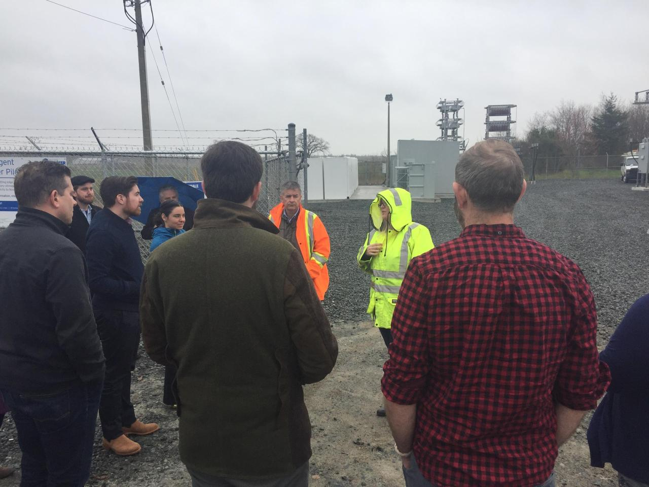 A group touring an electrical site with battery and grid equipment behind a fence.