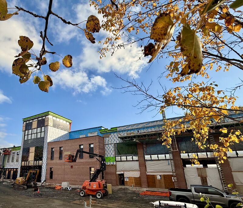 A building under construction with brickwork, exposed exterior insulation and construction equipment beneath autumn trees.
