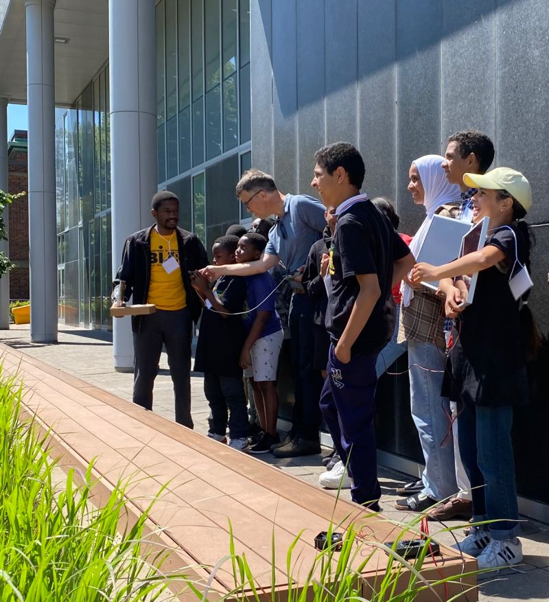 David Brushett demonstrating a small device to young participants outside a building.