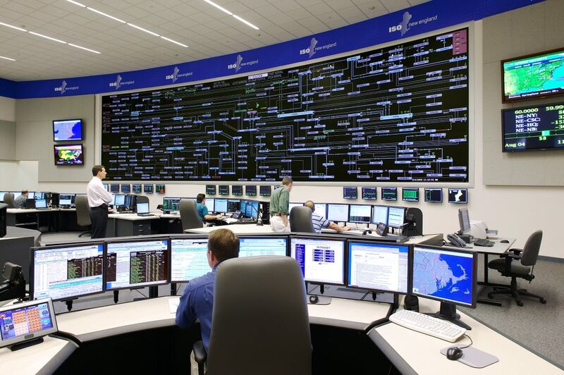 Operators at computer workstations beneath a large grid display in an ISO New England control room.