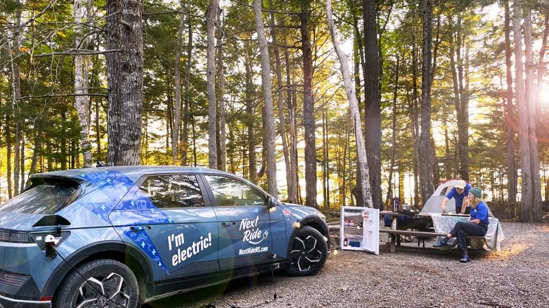A Next Ride electric vehicle at a wooded campsite beside a picnic table.